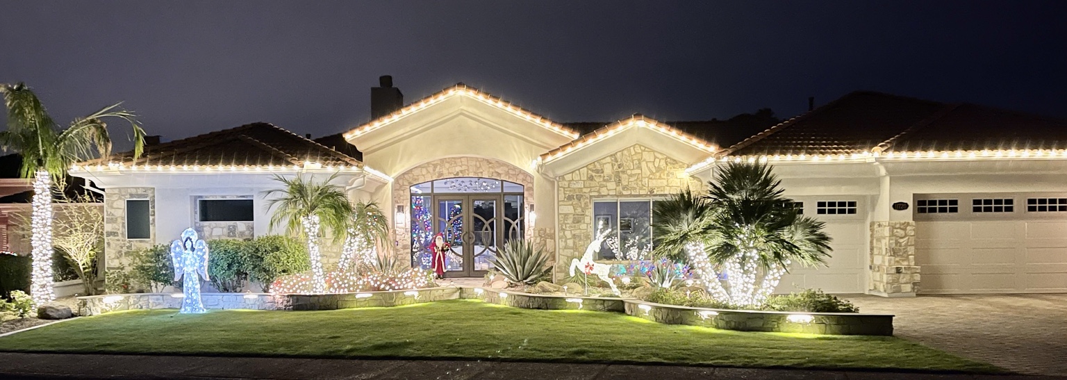 Completed outdoor Christmas display with warm white roofline lights, illuminated palms, shrubs, and holiday decorations