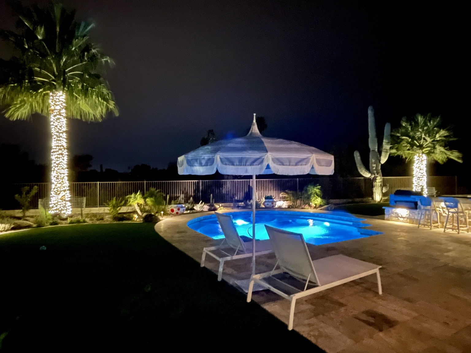Palm trees wrapped in warm white lights around a backyard swimming pool and patio at night